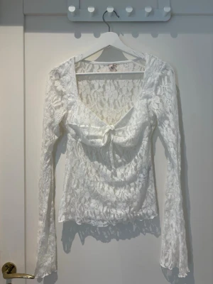 Vit spetsblus med knyt från Nelly - Supersöt vit långärmad blus från Nelly i spets med blommigt mönster och knyt framtill. Djup v-ringning och transparent material ger en trendig look. Perfekt att styla med jeans eller kjol för en cool outfit.
