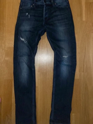 Mörkblå slim fit jeans med slitningar - Säljer ett par mörkblå slim fit jeans med snygga slitningar på låren och knäna. Jeansen har klassisk femficksmodell, knappgylf och lätt tvättad look. Perfekta för en avslappnad och trendig stil.