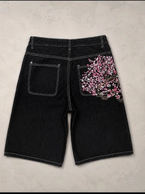 Svarta jeansshorts med broderat cherry blossom. - Unika svarta jeansshorts med vit kontrastsöm och ett broderat träd med rosa och vita blommor på bakfickan. Shortsens lösa passform och coola broderidetalj gör dem till ett statement-plagg för dig som vill sticka ut.
