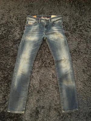 Blå slim fit jeans från Diesel - Säljer ett par blå slim fit jeans från Diesel med snyggt slitna detaljer och ljusa partier på låren och knäna. Klassisk femficksmodell med gulbruna sömmar och metallmärke vid fickan. Perfekta för en trendig och avslappnad stil. Storlek W30 och L32. Skriv till mig om du har några frågor.