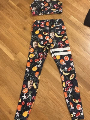 Färgglatt träningsset från Stronger - Säljer ett snyggt träningsset från Stronger i storlek S. Setet består av sport-bh och tights med färgglatt fruktmönster i orange, rött, gult och grönt på mörk botten. Materialet är 85% polyester och 15% elastan, vilket ger bra stretch och komfort.