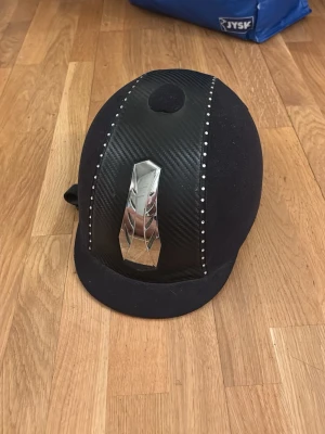 Svart ridhjälm med silverdetalj - Snygg svart ridhjälm från RIF med kolfiberliknande yta, silverfärgad detalj framtill och små glittriga stenar längs mitten. Hjälmen har en klassisk form och passar perfekt för dig som vill ha både stil och funktion i stallet.