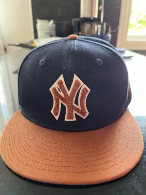 Marinblå och brun New Era keps - Snygg marinblå keps från New Era med platt, brun skärm och broderad NY-logga framtill i brunt och vitt. Klassisk snapback-modell med ventilationshål och kontrastfärgad knapp på toppen. Perfekt för dig som vill ha en stilren streetwear-look.storlek 57,7 cm