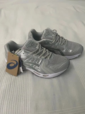 Gråa Asics sneakers med meshdetaljer -  Gråa sneakers från Asics med silvriga detaljer och meshpaneler för extra ventilation. Skorna har en sportig vibe med robust sula och klassisk snörning. Skriv om du har några frågor😊