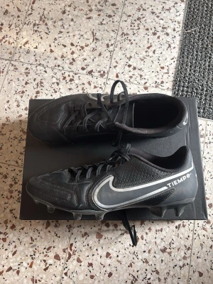 Nike Tiempo svarta fotbollsskor - Säljer ett par svarta Nike Tiempo fotbollsskor med vita detaljer och klassisk Swoosh-logga på sidan. Skorna har snörning, dobbar för gräsplan och ovandel i syntetmaterial med perforerade partier för bättre ventilation. Perfekta för dig som vill ha stilrena och funktionella skor på planen.