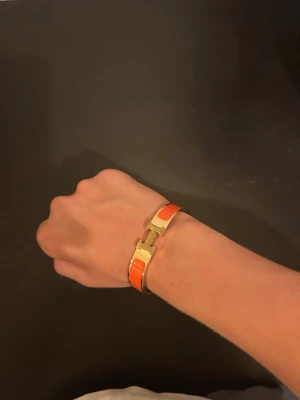 H armband - Snyggt armband i orange och guld med en stilren metallspänne i mitten. Armbandet har en blank finish och en modern, elegant design som sticker ut. Perfekt accessoar för att addera färg till din outfit.