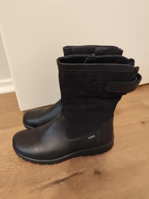 Svarta höga boots med dragkedja - Snygga svarta stövlar i en mix av läder och mocka. Modellen har en dekorativ rem med kardborre upptill och dragkedja på insidan för enkel på- och avtagning. Grov sula som ger bra grepp och en stilren look.