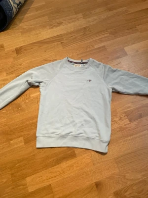 Ljusblå sweatshirt från GANT - Ljusblå sweatshirt från GANT med rund halsringning och diskret logga broderad på bröstet. Tröjan har långa ärmar, ribbade muddar vid ärmslut och nederkant samt en klassisk, avslappnad passform. Perfekt för en stilren och enkel look.
