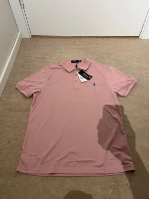 Rosa pikétröja från Polo Ralph Lauren - Säljer en snygg rosa pikétröja från Polo Ralph Lauren med klassisk krage och knappar framtill. Tröjan har en blå broderad logga på bröstet och är tillverkad i mjuk bomull. Perfekt för en clean och stilren look.