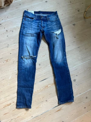 Blå slitna jeans från Hollister - Säljer ett par blå jeans från Hollister med slitningar och hål på båda benen. Jeansen har klassisk femficksmodell, normal midja och raka ben. Tillverkade i denim med snygg tvätt och detaljer som ger en cool, avslappnad look. Priset är ej skrivet i sten. 