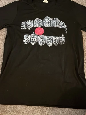 Svart t-shirt med tänder och smiley - Cool svart t-shirt med tryck av tänder med grills och en röd smiley mitt på. T-shirten har korta ärmar och en avslappnad passform. Perfekt för dig som vill sticka ut med en unik och edgy stil.