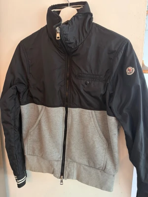Svart och grå vindjacka från Moncler - Snygg vindjacka från Moncler i svart och grått med hög krage och dragkedja framtill. Jackan har en bröstficka med knapp, sidofickor och Moncler-logga på ärmen. Överdel i glansig polyester och nederdel i mjuk bomullsmix. Stolek M passar S