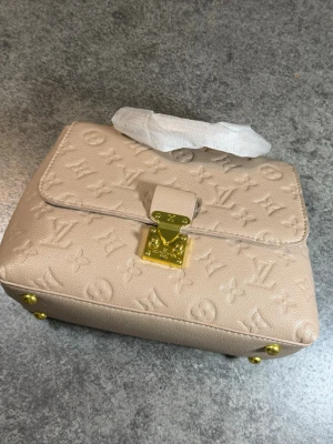 Beige handväska med monogram och gulddetaljer - Stilren beige handväska i präglat läder med klassiskt monogrammönster och guldfärgade metalldetaljer. Väskan har ett topphandtag och en elegant låsdetalj framtill. Perfekt accessoar för dig som gillar lyxig och tidlös design.