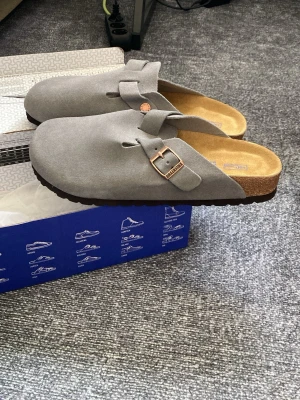 Grå Birkenstock tofflor med spänne - Säljer ett par gråa Birkenstock tofflor i mocka med klassiskt spänne i metall och bekväm korksula. Perfekta för dig som vill ha både stil och komfort hemma eller ute. Innersulan är mjuk och formar sig efter foten.