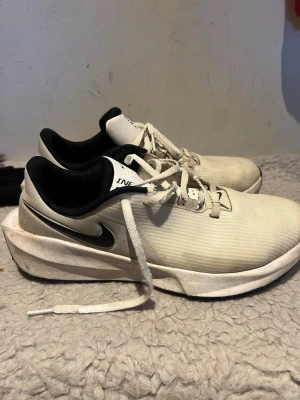 Vita Nike sneakers med svart sula - Säljer ett par vita Nike sneakers med svart sula och svart swoosh på sidan. Skorna har lågt skaft, vita snören och en slitstark yttersula med grovt mönster för bra grepp. Insidan är svart och ovandelen är i textilmaterial.