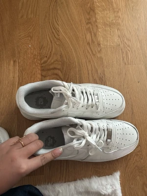 Vita Nike Air Force 1 sneakers - Klassiska vita Nike Air Force 1 sneakers med lågt skaft och perforerad tåbox. Skorna har vit sula, vita skosnören och den ikoniska swoosh-loggan på sidan. Tillverkade i läder med detaljer i metall på snörningen. Perfekta för en clean och stilren look.