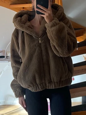 Brun teddy/pälsjacka från Zara - Mysig brun jacka från Zara med huva och dragkedja. Perfekt för kyliga dagar med sin fluffiga och varma design. Jackan har långa ärmar och en avslappnad passform. Passar xs/s beroende på hur man vill att den ska sitta då den är lite oversize. Bilderna ger ingen rättvis bild på färgen. Jackan är knappt använd. 