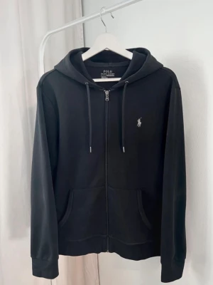 Svart hoodie från Polo Ralph Lauren - Snygg svart hoodie med dragkedja från Polo Ralph Lauren. Klassisk design med huva och dragsko, två fickor framtill och den ikoniska loggan broderad på bröstet. Tillverkad i mjukt bomullsmaterial som är skönt att bära.