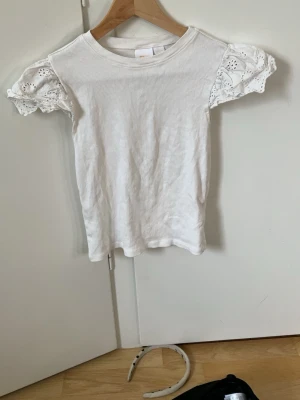 Vit topp med spetsärmar från Gina Tricot - Supersöt vit topp från Gina Tricot med korta puffärmar i spets och rund halsringning. Toppen är gjord i mjuk bomull och har en enkel, clean look med fina detaljer på ärmarna. Perfekt för dig som gillar en feminin och fräsch stil.