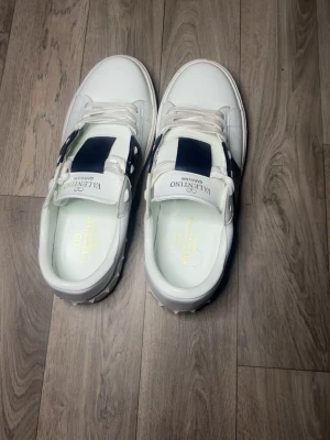 Vita sneakers från Valentino - Snygga vita sneakers från Valentino med bred svart detalj på sidan och vita skosnören. Skorna har en klassisk låg siluett och är tillverkade i slätt läder. Perfekta för dig som vill ha en stilren och exklusiv look.
