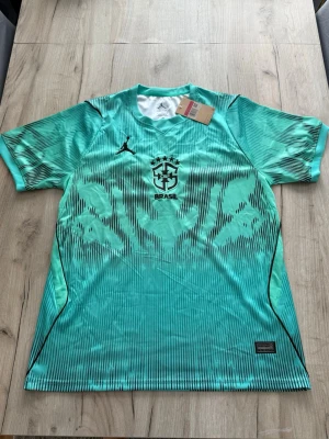 Turkos Brasil Dri-Fit fotbollströja - Snygg turkos fotbollströja från Jordan med Brasil-logga och fem stjärnor på bröstet. Tröjan har svart grafiskt mönster, rund hals och är tillverkad i lätt, snabbtorkande Dri-Fit-material som passar perfekt till träning eller match.