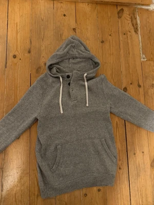 Stickad hoodie - grå hoodie med huva och vita dragsnören. Tröjan har knappslå vid halsen och en stor magficka framtill. Tillverkad i mjukt material och har ribbade muddar vid ärmslut och nederkant. Perfekt för chill dagar och enkel att matcha med jeans.