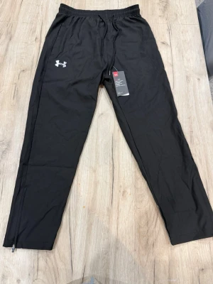 Svarta träningsbyxor Under Armour - Snygga svarta träningsbyxor från Under Armour med elastisk midja och dragkedjor vid bensluten. Byxorna har en diskret logga på låret och är tillverkade i ett lätt och funktionellt material som passar perfekt till träning eller sport.