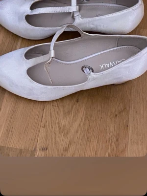 Vita ballerinaskor med T-rem - Säljer ett par vita ballerinaskor från Catwalk med klassisk T-rem och rundad tå. Skorna har en liten klack och spänne med dekorativ detalj. Materialet ser ut att vara konstläder med en matt finish. Perfekta för en stilren look.