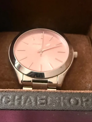 Silverfärgad klocka Michael Kors - Stilren klocka från Michael Kors med silverfärgat armband i metall och ljusrosa urtavla. Klassisk design med minimalistiska markeringar och rund boett. Perfekt accessoar för dig som gillar en clean look.