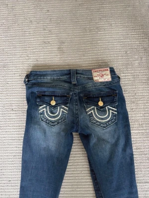 Blå bootcut jeans True Religion - Mörkblå True Religion Joey Low Rise jeans i storlek 25. Väldigt fina och i bra skick. Innerbenslängd cirka 79 cm och midjemått cirka 37 cm rakt över.