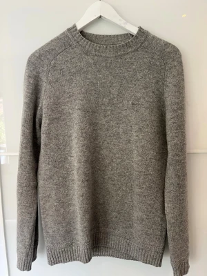 Grå stickad tröja från Gant - Klassisk grå stickad tröja från Gabr med diskret logga broderad på bröstet. Tröjan har rund halsringning, långa ärmar och ribbade muddar vid ärmslut och nederkant. Perfekt för hösten.