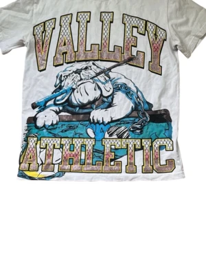 Vit t-shirt med VALLEY ATHLETIC-tryck - Cool vit t-shirt med stort färgglatt VALLEY ATHLETIC-tryck och en tecknad bulldogg med kedja på framsidan. T-shirten har korta ärmar och en avslappnad passform. Perfekt för dig som gillar streetwear och grafiska motiv.
