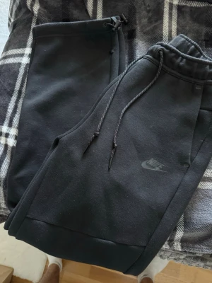 Nike Tech Fleece Raka byxor - Svarta mjukisbyxor från Nike med dragsko i midjan och diskret Nike-logga på benet. Byxorna har muddar vid bensluten och är gjorda i mjukt, fleeceliknande material. Perfekta för en avslappnad och sportig stil.