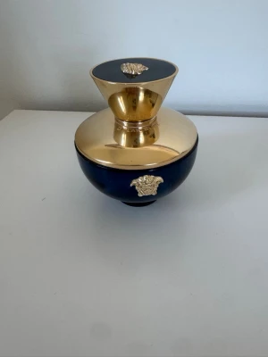 Versace Dylan Blue - Elegant parfymflaska i mörkblått glas med rund form och detaljer i blank guldmetall. Flaskan har ett exklusivt utseende med ett guldigt Medusahuvud på framsidan och en stor, konformad guldkork. Perfekt som lyxig inredningsdetalj eller för din favoritdoft. Parfymen är nästan full. Några repor på det blåa på flaskan. 