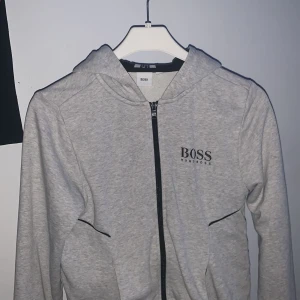 Grå hoodie från Boss - Säljer en stilren grå hoodie från Boss med dragkedja och svart logotyp på bröstet. Tröjan har en justerbar huva med svart foder och ribbade muddar vid ärmslut och nederkant. Perfekt för en avslappnad look. I storlek xxs
