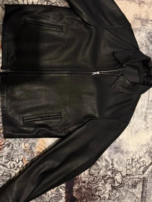 Svart skinnjacka från Jack & Jones, äkta läder - Snygg svart skinnjacka från Jack & Jones med klassisk krage och dragkedja framtill. Två raka fickor på sidorna och stilren design som passar till många olika outfits. Perfekt för dig som vill ha en tidlös och cool look.