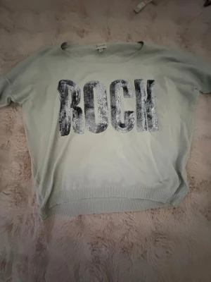 Ljusgrå tröja med paljetter ROCK - Trendig ljusgrå tröja med stora bokstäver 'ROCK' i silvriga paljetter på bröstet. Tröjan har rund halsringning, ribbad nederkant och är tillverkad i mjukt material. Perfekt för dig som vill sticka ut med en cool detalj.