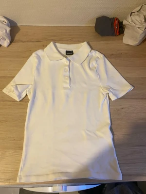 Vit kortärmad pikétopp - Klassisk vit pikétopp med krage och tre knappar framtill. Toppen har korta ärmar och är gjord i ett mjukt bomullsmaterial som känns skönt mot huden. Enkel och stilren design som passar till det mesta.
