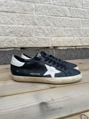 Golden Goose Super-Star svarta sneakers strl 42  - Svarta Golden Goose Super-Star sneakers i storlek 42 med vit stjärna och vit häl.  Använda men fortfarande i bra skick. Klassisk låg modell i läder.  Säljes eftersom storleken inte passar mig bra.