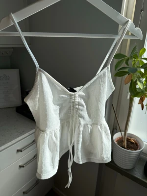 Linne - Söt vit peplumtopp från Gina Tricot i strukturerat tyg. Smala axelband, rynkad byst med knytning framtill och fin volang nertill. Perfekt till sommar, semester eller fest.  Storlek; S Skick; Ny med prislapp Material; 98% polyester, 2% elastan  #peplum #sommar #topp #ginatricot #scandi