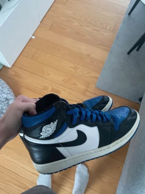 Svartvita och blå höga sneakers - Säljer ett par höga sneakers i svart, vitt och blått med blå skosnören och blå yttersula. Skorna har en klassisk siluett med tydlig swoosh på sidan och Air Jordan-logga vid ankeln. Materialet är en mix av läder och syntet.