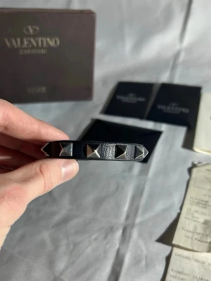 Valentino svart läderarmband - Snyggt svart armband från Valentino i äkta läder med silverfärgade pyramidnitar runt om. Armbandet har ett stilrent och edgy uttryck, perfekt för dig som gillar att sticka ut med accessoarer. Justerbart spänne för bästa passform. I slutet är lädret slitet men men kan enkelt suprlimma.. orgional box, extra nitar och kvitto medföljer 