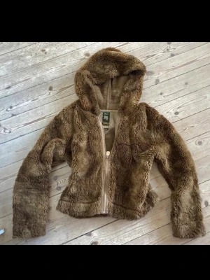 Brun teddyjacka från Miss Sixty - Säljer en fluffig brun teddyjacka från Miss Sixty med dragkedja och huva. Jackan har en croppad passform och är gjord i mjukt, fuskpälsliknande material som ger en riktigt cozy vibe. Perfekt för dig som vill ha en statementjacka till hösten eller vintern.