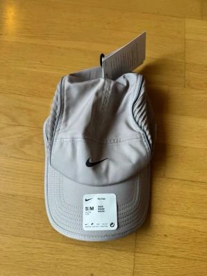 Ljusgrå Nike keps S/M - ljusgrå löparkeps från Nike med klassisk böjd skärm och svart Nike-logga framtill. Tillverkad i lätt och ventilerande material, perfekt för träning eller vardagsstil. Justerbar passform och sportig design.