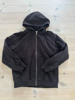Svart zip hoodie med huva Weekday - Klassisk svart hoodie med dragkedja framtill och stor ficka på magen. Tröjan har huva med dragsko och ribbade muddar vid ärmar och nederkant. Perfekt för en avslappnad och stilren look.