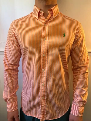 Orangevit randig skjorta från polo Ralph Lauren  - Snygg och stilren skjorta från polo Ralph Lauren  med orange och vita ränder. Perfekt för dig som gillar stilrena detaljer och en fräsch look.