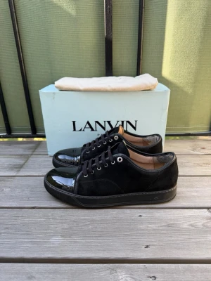 Svarta sneakers från Lanvin - Snygga svarta sneakers från Lanvin med ovandel i mocka och lackade tåpartier. Skorna har svarta skosnören, metallöljetter och en klassisk låg siluett. Perfekta för dig som gillar stilrena och exklusiva detaljer. Allt på bilden ingår. Finns kvar beläggning på hälen men märks inte vid användning