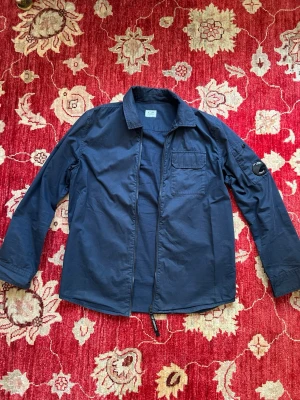 Marinblå overshirt från CP Company - Snygg marinblå overshirt från CP Company med dragkedja, bröstficka och klassisk patch på ärmen. Tillverkad i ett lätt bomullstyg som passar perfekt för lager-på-lager. Enkel och stilren design med diskreta detaljer.