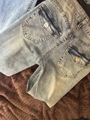 Ljusblåa jeans  - Säljer ett par ljusblå jeans med snygga och unika bakfickor med dekorativa sömmar och knappar. med knappgylf och bälteshällor. Perfekta snygga med diamanter 