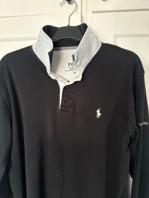 Ralph Lauren långärmad piketröja - Säljer en svart pikétröja från Polo Ralph Lauren. Riktigt stilren och extremt skön att bära. Stilren och funkar i princip alla sammanträdanden. Storlek M. Vet ej nypris
